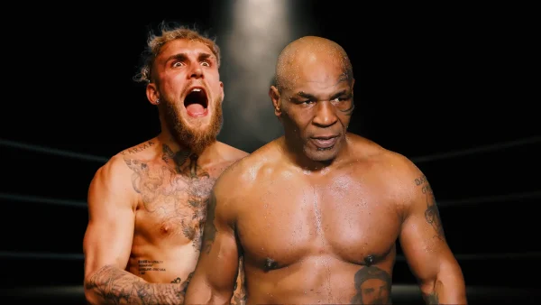 An image featuring Mike Tyson to the immediate front and Jake Paul to the side both shirtless against a black background under a spotlight.